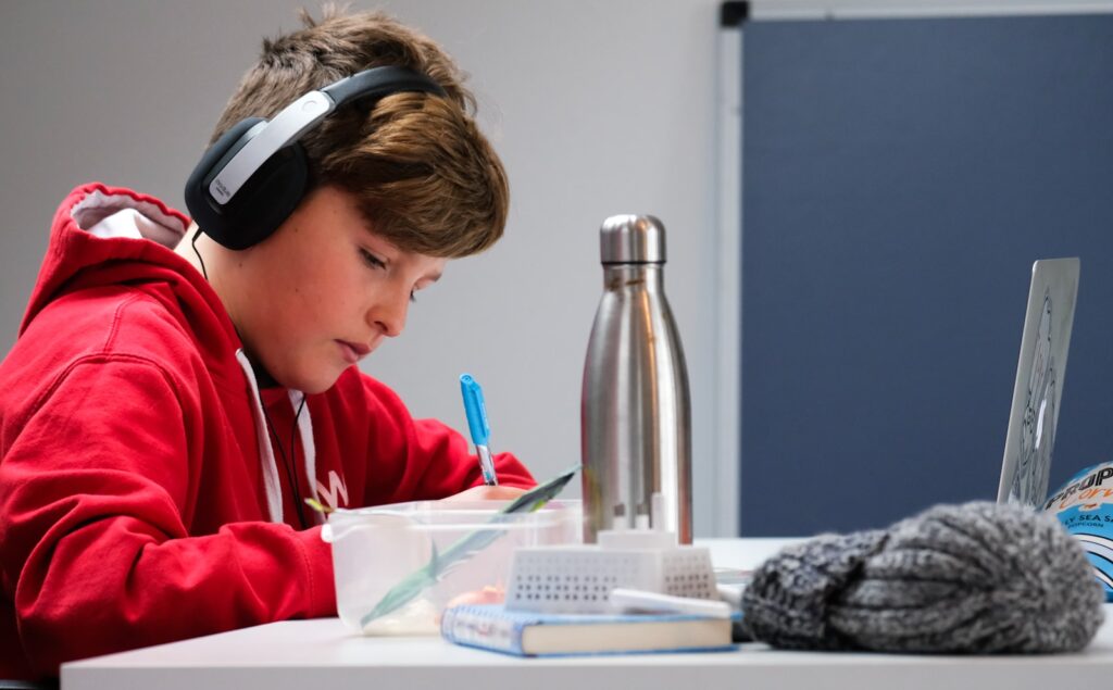 child in a red hoody using a laptop and headphones to receive online tutoring to help bring him up to speed at school